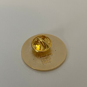 May include: A gold-colored round pin with a pin back. The back of the pin has the text "MUCHO LOVE PINS" stamped into the metal. The pin is likely a collectible or decorative item.