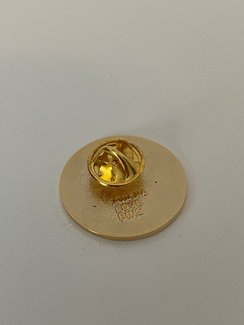 May include: A gold-colored round pin with a pin back. The back of the pin has the text "MUCHO LOVE PINS" stamped into the metal. The pin is likely a collectible or decorative item.
