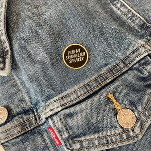 May include: A round, black and gold enamel pin with the text "FLUENT SPANGLISH SPEAKER" in white lettering. The pin is attached to a blue denim jacket.