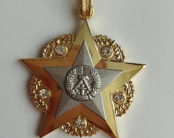 May include: A gold and silver five-pointed star pendant with a hammer and compass emblem in the center. The star is surrounded by a decorative border with rhinestones.