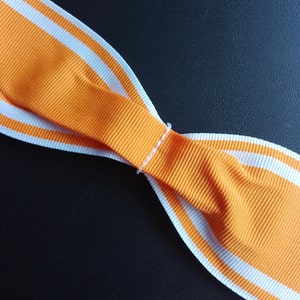May include: Orange ribbon with two white stripes, folded in the shape of a bow.