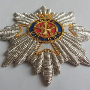May include: A silver and gold embroidered badge with a red, white, and blue circular design in the center. The center design features a crown, a letter "R", and the word "VICTORIA".