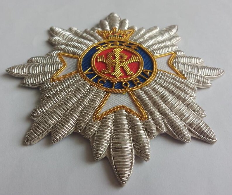 May include: A silver and gold embroidered badge with a red, white, and blue circular design in the center. The center design features a crown, a letter "R", and the word "VICTORIA".
