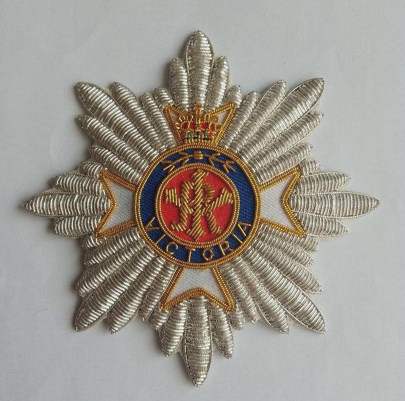 May include: A silver and gold embroidered badge with a red and blue circle in the center. The circle features a crown and the word "VICTORIA".