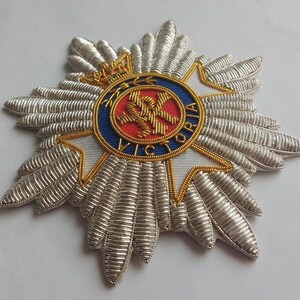 May include: A silver embroidered badge with a red, white, and blue circular emblem in the center. The emblem features a crown, a cross, and the text "VICTORIA".