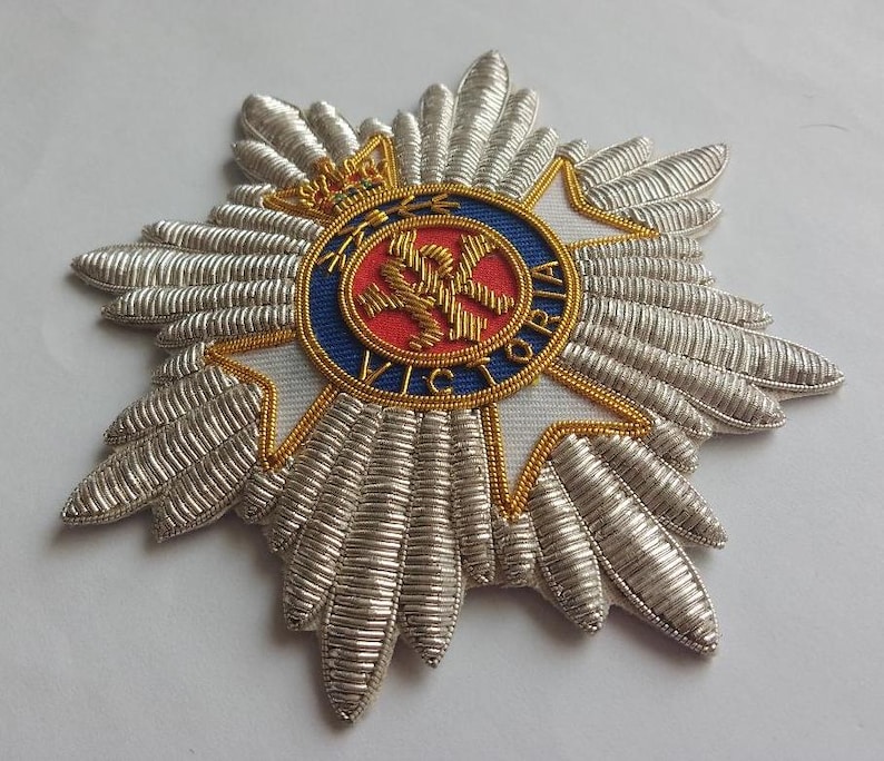 May include: A silver embroidered badge with a red, white, and blue circular emblem in the center. The emblem features a crown, a cross, and the text "VICTORIA".