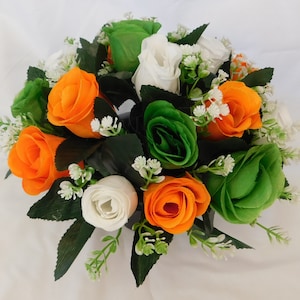 May include: A floral arrangement featuring orange, white and green roses. The roses are arranged in a circle and are surrounded by small white flowers.