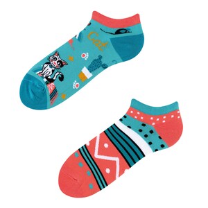 Può includere: Un paio di calzini corti colorati con un design a gatto su una calza e un motivo geometrico sull'altra. I calzini sono turchese, rosa e bianco.