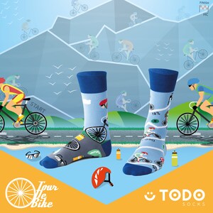 May include: A pair of blue and gray socks with a cycling theme. The socks feature a cyclist riding a bike, a mountain range, and the words "Tour de Bike". The socks are on a blue and yellow background with a pair of sunglasses, a water bottle, and a helmet.