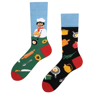 Könnte beinhalten: Ein Paar bunte Socken mit einem Koch- und Küchendesign. Eine Socke zeigt einen Koch, der einen Teller mit Essen hält, während die andere Socke eine Vielzahl von Küchenutensilien und Zutaten zeigt.