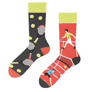 Può includere: Un paio di calzini colorati a tema tennis. Un calzino presenta un giocatore di tennis su un campo di terra rossa con una rete e una racchetta da tennis. L'altro calzino presenta uno sfondo nero con racchette da tennis e palline da tennis.
