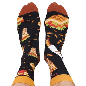 Könnte beinhalten: Schwarze Socken mit einem sich wiederholenden Muster aus Sandwiches, Pommes Frites und Gewürzen. Die Socken haben braune Fersen und Zehen sowie gelbe Bündchen. Ein lustiger, von Lebensmitteln inspiriertes Novelty-Item.