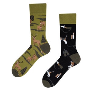 Könnte beinhalten: Ein Paar Socken mit Jagdmotiv. Eine Socke hat einen grünen Hintergrund mit Hirschen und Bäumen, während die andere Socke einen schwarzen Hintergrund mit einem Jagdhund, einer Waffe und anderer Jagdausrüstung hat.