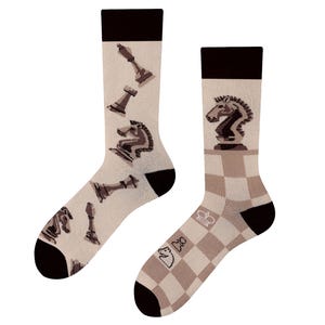 Könnte beinhalten: Ein Paar beigefarbene Socken mit einem Schachthema. Eine Socke hat ein Schachbrettmuster und die andere Socke hat ein Muster aus Schachfiguren, darunter ein Springer, ein Turm und ein Bauer.