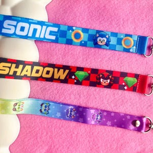 May include: Three colourful lanyards featuring characters and logos from the Sonic the Hedgehog video game series. The lanyards are blue, red, and purple, with the names "SONIC" and "SHADOW" printed on them. Each lanyard has a metal clasp.