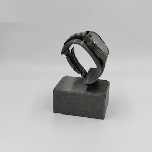 Könnte beinhalten: Eine schwarze Uhr mit silbernem Zifferblatt und schwarzem Armband wird auf einem schwarzen Ständer präsentiert.
