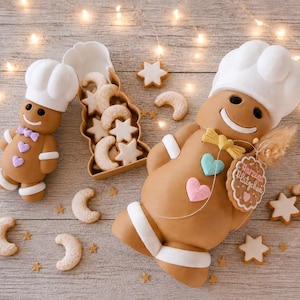 Könnte beinhalten: Zwei Lebkuchenfiguren mit weißen Kochmützen, eine kleine und eine große. Die große Figur trägt eine gelbe Fliege, ein türkisfarbenes Herz, ein rosa Herz und ein rundes Schild mit der Aufschrift "Herzens Plätzchen". Ein Behälter enthält Kekssterne.
