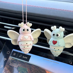 Könnte beinhalten: Zwei bienenförmige Auto-Lufterfrischer, die an einer Schnur hängen. Einer ist hellrosa mit einem weißen Herz, der andere hellblau mit einem roten Herz. Beide haben große Wackelaugen, herzförmige Fühler und cremefarbene Flügel mit kleinen Löchern.