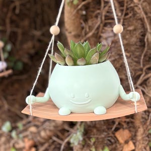 Könnte beinhalten: Ein hellgrüner Keramiktopf in Form eines lächelnden Gesichts mit Armen und Beinen, der eine Sukkulente beherbergt. Der Topf hängt an einer Holzschaukel mit Schnur.