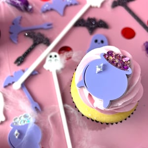 May include: A purple cauldron cupcake topper with rhinestones sits on top of a pink and white frosted cupcake. The cupcake is on a pink surface with other Halloween themed decorations.