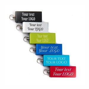 Könnte beinhalten: Eine Auswahl an rechteckigen Schlüsselanhängern in verschiedenen Farben: Schwarz, Silber, Grün, Blau und Rot. Jeder Schlüsselanhänger hat einen silbernen Ring und den weißen Text "Your text Your LOGO", ideal für die Personalisierung.
