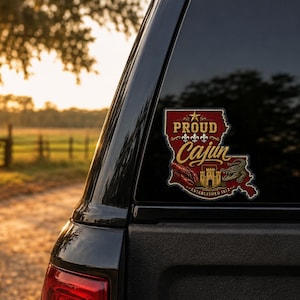 May include: A decal shaped like the state of Louisiana in red and gold, featuring the text PROUD Cajun, three white fleur-de-lis, a crawfish, an alligator, and a castle illustration with the text ESTABLISHED 1812, applied to a vehicle window.