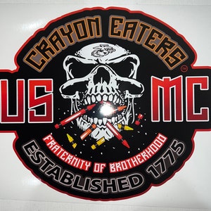 May include: A black and red circular sticker with a skull wearing a USMC hat and biting down on crayons. The text reads "Crayon Eaters MC" and "Fraternity of Brotherhood Established 1775".