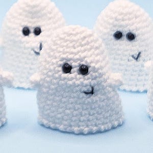 Può includere: Sei fantasmini bianchi all'uncinetto fatti a mano, con occhi di perline nere e sorrisi ricamati, su una superficie azzurra. L'immagine include il testo "papesa" e un logo.