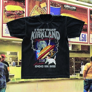 Può includere: T-shirt nero con la scritta "I GOT THAT KIRKLAND Signature DOG IN ME". La grafica include un hot dog, una bibita e due cani. La maglietta è esposta in un ambiente di ristorazione.