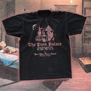 Könnte beinhalten: Schwarzes T-Shirt mit einem rosa Aufdruck eines Hauses und dem Text "The Pink Palace Apartments". Das T-Shirt enthält auch den Satz "Your Other Home Awaits" und den Ort "Hubland, Oregon."