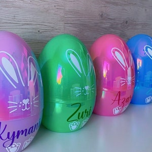 May include: Four large, iridescent Easter egg containers in a row. The eggs are in shades of purple, green, pink, and blue. Each egg has a white bunny face design and a name in cursive. The eggs are sitting on a white surface.