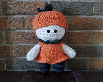 May include: A crocheted pumpkin doll wearing an orange dress and a black and orange hat. The doll is standing on a brown wooden surface.