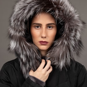 May include: A black winter coat with a large hood trimmed with silver and brown fur. The coat has a quilted design and a dark gray lining at the cuffs. The person is wearing dark lipstick.