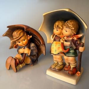 Puede incluir: Dos figuras de cerámica que representan niños bajo paraguas. Una figura muestra a un niño sentado bajo un paraguas marrón, mientras que la otra muestra a dos niños de pie juntos bajo un paraguas grande y claro, vistiendo ropa tradicional.