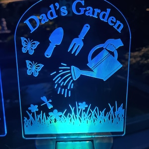 May include: A blue LED garden stake with the words "Dad's Garden" at the top. The stake features garden-themed illustrations, including butterflies, a trowel, a fork, a watering can, and flowers. The stake is solar-powered.