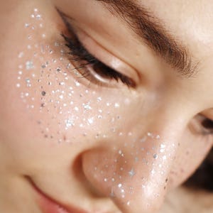 Puede incluir: Primer plano del rostro de una persona con purpurina plateada aplicada en la piel alrededor del ojo y la mejilla. La persona tiene delineador de ojos oscuro y un color de labios rosa claro. Se ve un pendiente floral blanco.