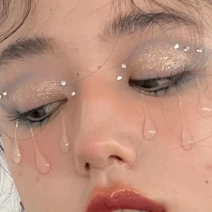 Op de afbeelding: Een make-up look met glinsterende gouden oogschaduw op de oogleden, geaccentueerd met kleine doorzichtige strass-steentjes langs de arcadeboog en wimperlijn. Doorzichtige, traanvormige versieringen zijn onder de ogen aangebracht.