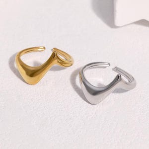 Pode incluir: Dois anéis abertos com um design ondulado em forma de V. Um anel tem um acabamento em tom dourado polido, enquanto o outro tem um acabamento em tom prateado polido. Ambos os anéis apresentam uma banda fluida e contínua que afunila numa ponta.