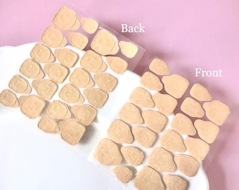 Può includere: Due fogli trasparenti contenenti diversi piccoli cerotti adesivi color beige. Il foglio a sinistra è etichettato Back e quello a destra Front. Ogni cerotto ha una forma arrotondata e irregolare con un'indentazione rettangolare.