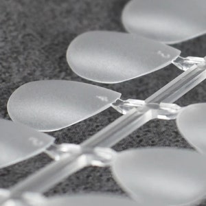 May include: A set of clear, frosted plastic nail art display tips arranged in a row on a central connecting stem. Each individual tip has a rounded, teardrop-like shape with a smooth, matte finish, designed for showcasing various nail polish colors.