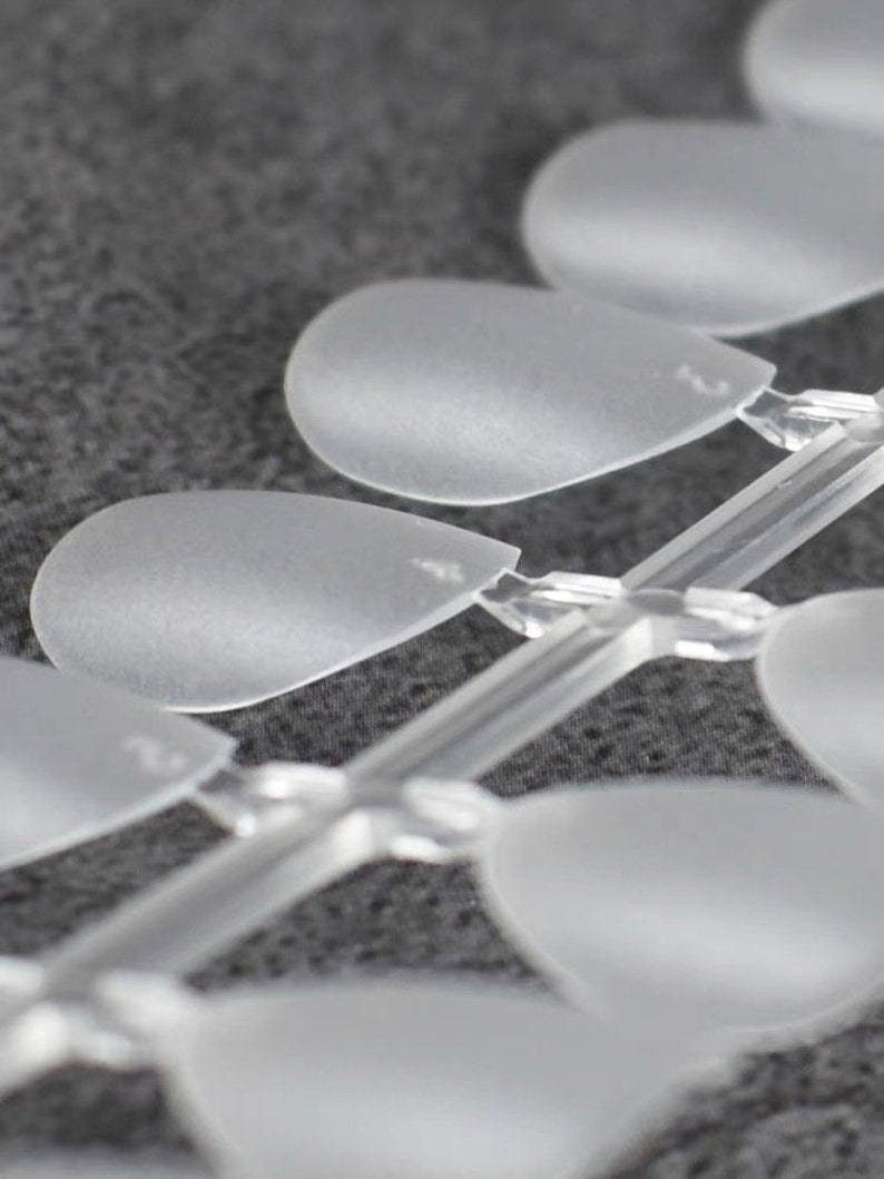 May include: A set of clear, frosted plastic nail art display tips arranged in a row on a central connecting stem. Each individual tip has a rounded, teardrop-like shape with a smooth, matte finish, designed for showcasing various nail polish colors.