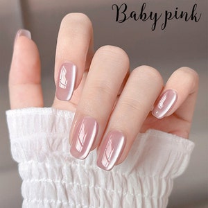 Puede incluir: Una mano con esmalte de u&ntilde;as rosa beb&eacute;. Las u&ntilde;as son de un rosa suave y apagado con un acabado brillante. El texto "Baby pink" est&aacute; escrito en cursiva en la esquina superior derecha. La mano lleva una manga blanca texturizada.