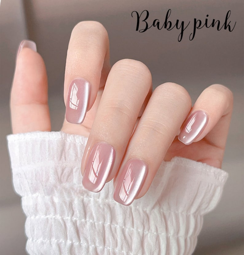Puede incluir: Una mano con esmalte de u&ntilde;as rosa beb&eacute;. Las u&ntilde;as son de un rosa suave y apagado con un acabado brillante. El texto "Baby pink" est&aacute; escrito en cursiva en la esquina superior derecha. La mano lleva una manga blanca texturizada.