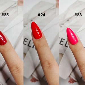 May include: Three almond-shaped fingernails with glossy polish in shades of deep red, bright red, and hot pink, labeled #25, #24, and #23 respectively. Each nail displays a smooth, reflective finish with a distinct color variation across the set.