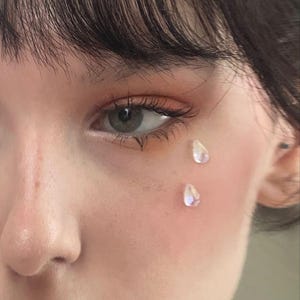 May include: Close-up of an eye with long eyelashes and a subtle orange eyeshadow. Two iridescent, teardrop-shaped gems are adhered to the cheek. The skin has a soft, pinkish hue.
