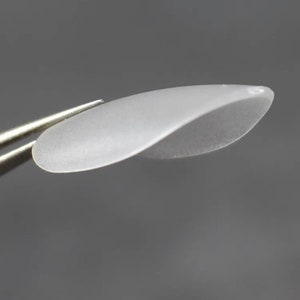 May include: A small, translucent, frosted white petal-shaped component with a smooth, curved surface. The object has a delicate, elongated oval form with a slight twist, featuring a small hole at one end for attachment purposes.