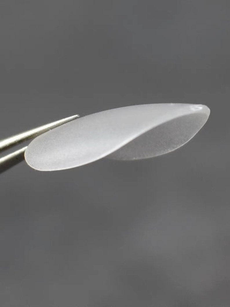 May include: A small, translucent, frosted white petal-shaped component with a smooth, curved surface. The object has a delicate, elongated oval form with a slight twist, featuring a small hole at one end for attachment purposes.