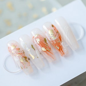 May include: Set of six long, clear acrylic false nails with a peach and white marbled design and gold flakes.