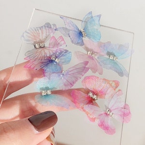 May include: Assortment of iridescent butterfly embellishments with rhinestones. The butterflies are in shades of pink, blue, and purple.