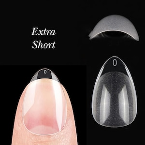 May include: Clear, extra short, oval-shaped nail tips are displayed against a black background. One tip is adhered to a finger, showcasing the transparent design. The text "Extra Short" is visible in the image.